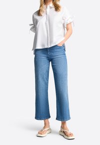 Donna con una blusa bianca a maniche corte con maniche annodate, jeans blu a vita alta e sandali platform avorio, in piedi con una mano in tasca.