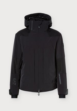 Un homme se tient face à l’objectif, vêtu d’une veste noire à capuche, d’un pantalon olive et de bottes noires, sur un fond clair uni.