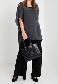 Luźna szara bluzka z wycięciami na ramionach i koralikowym dekoltem, zestawiona z czarnymi spodniami o szerokich nogawkach i czarną, strukturalną torebką.