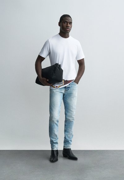 Άνδρας με λευκό μπλουζάκι, ανοιχτό μπλε τζιν, μαύρα μποτάκια, κρατώντας μια διπλωμένη μαύρη ζακέτα μπροστά σε απλό ανοιχτό γκρι φόντο.