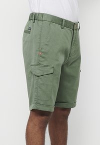 Man som bär olivgrön cargoshorts med uppvikta nederkanter och flera fickor, i kombination med en vit t-shirt, visad från midjan till knäna.