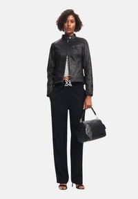 Veste en cuir noir avec un col haut, associée à un pantalon noir. Le modèle tient un sac à main noir avec un accent décoratif.