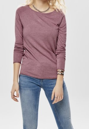 Camisa de manga larga burdeos con cuello estilo crew y una textura ligera, combinada con jeans ajustados azules y acentos de pulsera de cadena plateada.