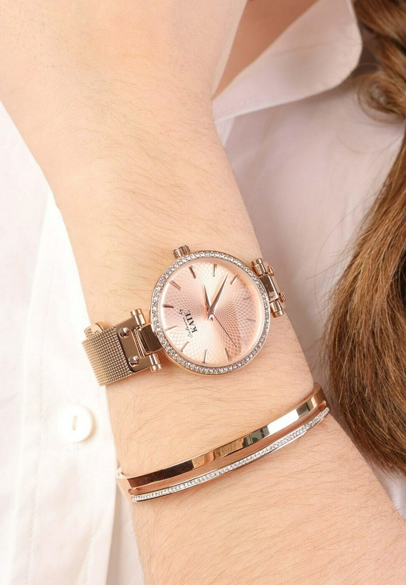 Roze-gouden polshorloge met een gestructureerde, lichtroze wijzerplaat en glinsterende accenten. Voorzien van een meshband en een slanke, gepolijste armband ernaast.