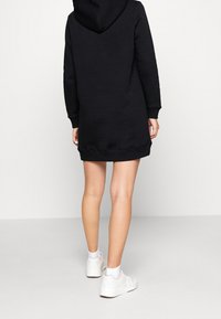 Robe sweat à capuche noire avec manches longues, poignets côtelés et coupe décontractée, associée à des baskets blanches. Texture lisse et longueur au genou.