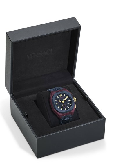 Montre Versace dans un boîtier octagonal bordeaux avec des accents dorés, cadran noir et bracelet texturé bleu, à l'intérieur d'une boîte noire avec un revêtement doux.