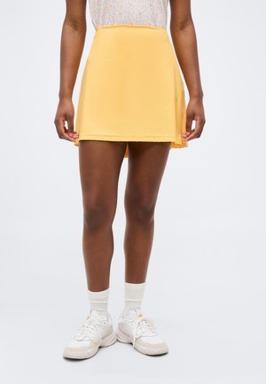 Γυναίκα που φοράει κίτρινη φούστα τένις, λευκό τοπ με σχέδια, λευκές κάλτσες και λευκά αθλητικά παπούτσια, στέκεται μπροστά σε απλό φόντο.