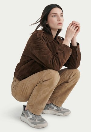 Woman squatting, wearing a brown suede jacket, light brown pants, and gray athletic sneakers, looking slightly upward with hands clasped.