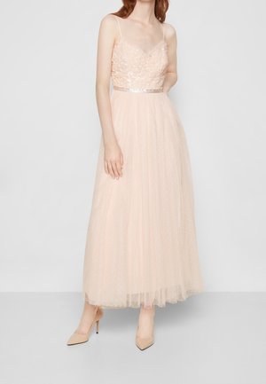 Kobieta w sukience z bezrękawnym, różowawym, cekinowym gorsetem i długą tiulową spódnicą, nosząca beżowe buty na szpic, stojąca na tle jednolitego tła.