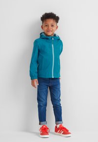 Teal windbreaker with a hood, front zipper, and elastic cuffs. Worn over blue jeans and red athletic shoes. Simple design, lightweight material.
