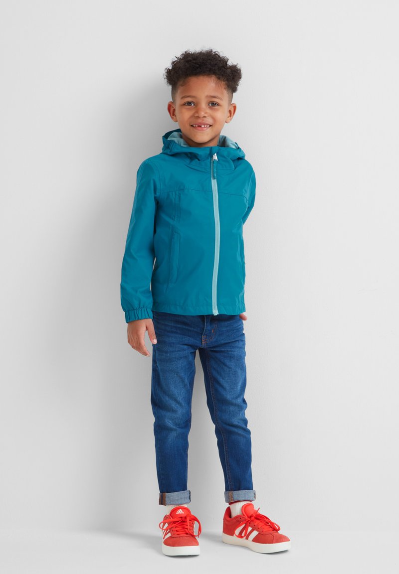 Teal windbreaker with a hood, front zipper, and elastic cuffs. Worn over blue jeans and red athletic shoes. Simple design, lightweight material.