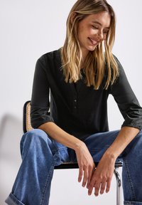 Femme souriante aux longs cheveux blonds assise sur une chaise, les jambes écartées, les mains reposant entre les genoux, portant un haut noir et un jean bleu.