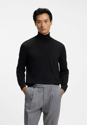 Uomo con maglione nero a collo alto e pantaloni grigi plissettati, in piedi con le mani in tasca davanti a uno sfondo bianco uniforme.