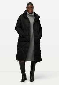 Abrigo de plumón negro con acolchado en diamante, cuello alto y bolsillos laterales, llevado sobre un vestido de cuello alto gris y combinado con botas negras hasta la rodilla.
