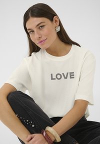 Bijela majica kratkih rukava s natpisom "LOVE" ispisanim crnim točkicama, u kombinaciji s tamnim trapericama ukrašenim rhinestonima i složenim narukvicama.