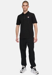 Svart randig polo med krage, med en liten logotyp och ihop med svarta byxor och vita sneakers.
