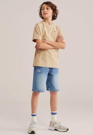 Bernas su garbanotais plaukais stovi sukryžiavęs rankas, dėvi smėlinės spalvos marškinėlius, džinsinius šortus, baltas kojines su mėlynomis juostelėmis ir smėlinės spalvos sportbačius.