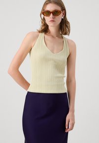 Gul ribbet tanktop med V-hals og halterneck-design, kombinert med en mørk lilla skjørt. Tilbehør inkluderer skjoldpadde-solbriller og øredobber.