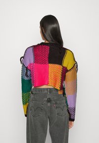 Gehaakte trui met een patchworkontwerp in levendige kleuren. Heeft een cropped snit, open gebreide textuur en gevlochten accenten. Gecombineerd met grijze jeans.