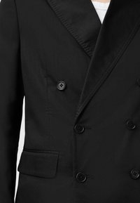 Blazer doppiopetto nero realizzato in tessuto testurizzato, con grandi bottoni neri e colletto a punta. Include una tasca laterale.