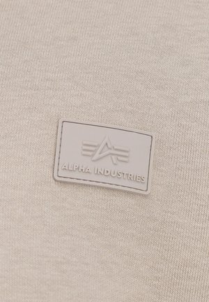 Béžová tkanina s textúrovaným pletením. Obsahuje šedú, obdĺžnikovú gumovú nášivku s embosovaným logom a dizajnom "Alpha Industries".