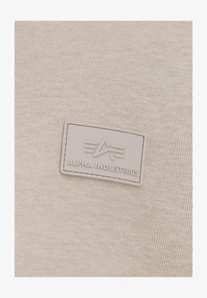 Béžová tkanina s textúrovaným pletením. Obsahuje šedú, obdĺžnikovú gumovú nášivku s embosovaným logom a dizajnom "Alpha Industries".