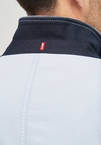 Světle modrá polo košile s námořnickým límcem, zdobená bílým prošíváním a malou červenou akcentní nášivkou na zadní straně. Viditelné detaily texturované tkaniny.
