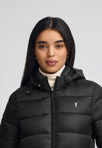 Czarna kurtka puchowa z zamkiem z przodu, regulowanym kapturem i poziomym pikowaniem. Posiada małe białe logo w okolicy klatki piersiowej.