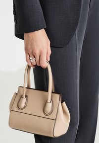 Una mano che tiene una piccola borsa in pelle beige, indossando un completo grigio scuro e anelli d'argento sulle dita, su uno sfondo semplice.