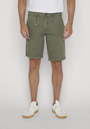 Mężczyzna w oliwkowozielonych teksturowanych szortach z podwiniętymi nogawkami i białych trampkach, stojący na tle gładkiego tła.