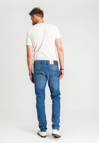 Mężczyzna w białej koszulce, niebieskich dżinsach z widocznymi tylnymi kieszeniami i białych trampkach, stojący na tle jednolitego jasnego tła.