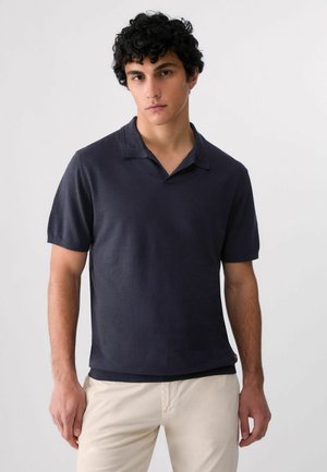 Giovane uomo con capelli ricci che indossa una polo blu scuro a maniche corte e pantaloni beige chiaro su uno sfondo chiaro semplice.