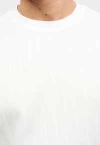 Κοντινό πλάνο ενός ατόμου που φοράει απλό λευκό μπλουζάκι με λαιμόκοψη ριμπ, εμφανίζοντας τον λαιμό και το πάνω μέρος του στήθους.