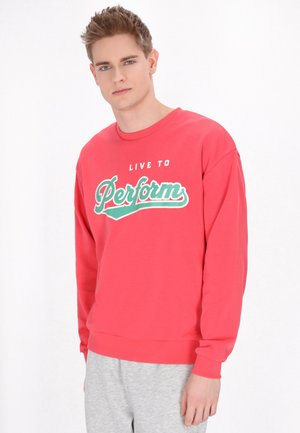 Rød bomuldssweatshirt med rund hals, lange ærmer og et trykt slogan i hvid og teal. Båret med grå sweatpants.