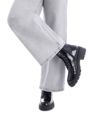 Botas de tobillo de charol negro con suela gruesa, que cuentan con cordones y una textura brillante, llevadas con jeans de pierna ancha grises.