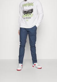 White long-sleeve shirt with black and neon green graphic design, paired with blue slim-fit pants and white sneakers with red and blue accents.