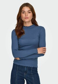 Blå ribbad långärmad topp med hög, volangkrage och vågiga manschetter. Bärs med mörka denimjeans som har vertikal sömnad.