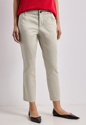 Personne portant un pantalon beige court, un pull rouge et des ballerines noires pointues debout sur un sol blanc contre un mur uni.