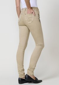 Jeans beige ajustados con una textura suave y bolsillos traseros, con costuras decorativas. Llevados con zapatos planos negros.