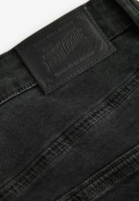 Crni traper s teksturiranom površinom, s kožnom etiketom pri struku koja glasi "Signature Denim Goods."