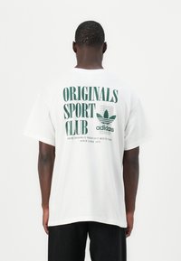 Tricou alb din bumbac cu text imprimat verde pe spate, incluzând "ORIGINALS SPORT CLUB" și logo-ul Adidas, cu mâneci scurte.
