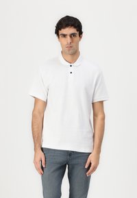 Hvid polo skjorte med klassisk krave, tre sorte knapper, korte ærmer og struktureret stof. Båret med blå jeans.