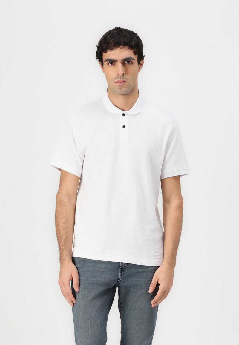 Hvid polo skjorte med klassisk krave, tre sorte knapper, korte ærmer og struktureret stof. Båret med blå jeans.