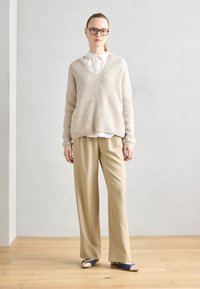 Pull en tricot beige avec un décolleté en V profond, superposé sur une chemise blanche à col. Assorti à un pantalon large beige clair et des chaussures bicolores.