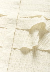 Teksturirana off-white tkanina s naboranim, slojevitim dizajnom. Ima suptilan uzorak i nježne, tekuće rubove. Mekan i lagan izgled.