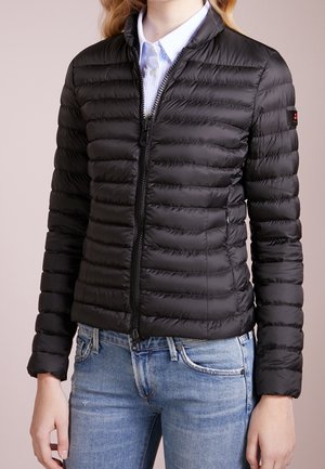 Zwarte gewatteerde donzen jas met ritssluiting aan de voorkant, gedragen over een lichtblauw overhemd met kraag en een lichtblauwe spijkerbroek.