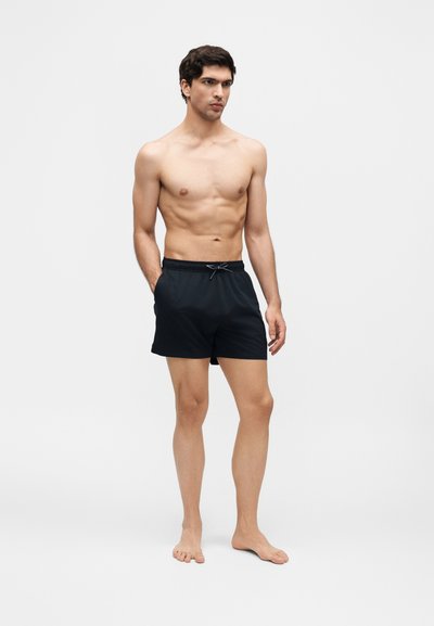 Άνδρας χωρίς μπλούζα, όρθιος ξυπόλυτος, φοράει μαύρο μαγιό με κορδόνι σε απλό ανοιχτό φόντο.