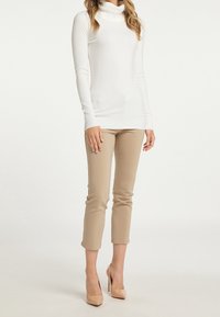 Witte coltrui, slanke pasvorm, geribbelde textuur. Beige cropped broek past strak. Model draagt naaktkleurige hoge hakken en poseert met één hand op de heup.