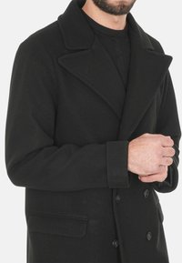 Cappotto in lana nera con colletto a picco, design doppiopetto e due tasche laterali. La trama appare liscia e calda.