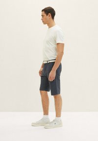 Männliches Outfit mit einem taillierten weißen T-Shirt, dunklen gemusterten Shorts und weißen Sneakers mit strukturierten Sohlen. Der Gürtel ist hell.